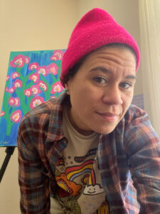 Close up photo of Lydia wearing a red beanie, a t-shirt, and a flannel shirt over top. A painting with a blue background and pink flowers can partially be seen in the background.