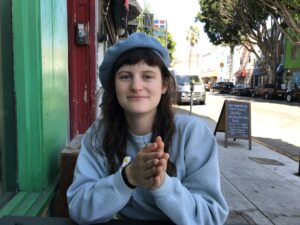 Photo of Zoe sitting outside in an urban setting, wearing a blue beret and light blue sweatshirt with hands clasped together, smiling slightly. 